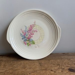 May include: White ceramic serving plate with a floral design featuring pink and blue flowers. The plate has a scalloped edge and a raised rim. The center of the plate features a yellow and white checkered pattern. The plate is marked "Made in England" and "Ryde, Isle of Wight".