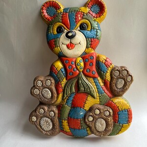 May include: A colorful, patchwork-style teddy bear wall hanging. The bear features a red bow tie with blue polka dots, and is made of various colors including red, blue, yellow, and green. The bear's paws and face are a light brown color.