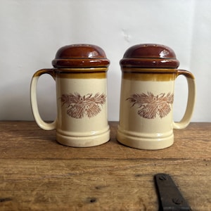 May include: A pair of ceramic salt and pepper shakers shaped like miniature beer steins. Each shaker features a cream-colored body with a brown rim, a brown handle, and a brown lid with small holes. A brown wheat sheaf graphic is on the front.