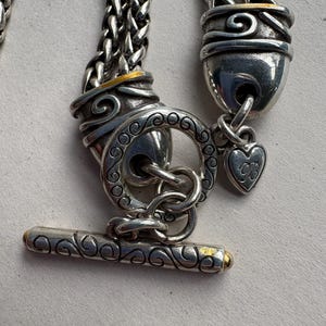 Pu&ograve; includere: Primo piano di una chiusura per collana argentata con chiusura a toggle. La chiusura presenta un'intricata lavorazione a volute e dettagli dorati. Un piccolo ciondolo a forma di cuore pende dalla chiusura. La catena &egrave; un design intrecciato.
