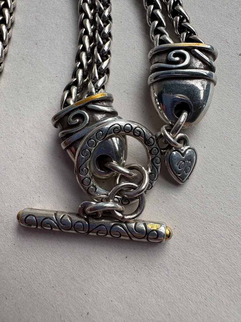 Pu&ograve; includere: Primo piano di una chiusura per collana argentata con chiusura a toggle. La chiusura presenta un'intricata lavorazione a volute e dettagli dorati. Un piccolo ciondolo a forma di cuore pende dalla chiusura. La catena &egrave; un design intrecciato.