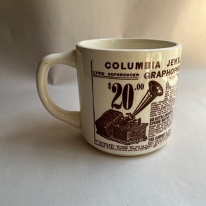 May include: White ceramic coffee mug with a brown illustration of a Columbia Jewel Lyric Reproducer Graphophone. The text "$20.00" is printed on the mug.