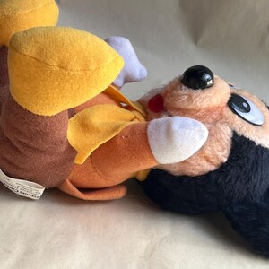 Op de afbeelding: Een pluche speelgoed met een stripfiguur met een zwarte kop, een oranje lichaam en gele accenten. Het speelgoed heeft een zwarte neus, grote ogen en een rode mond. Het speelgoed ligt op zijn zij.