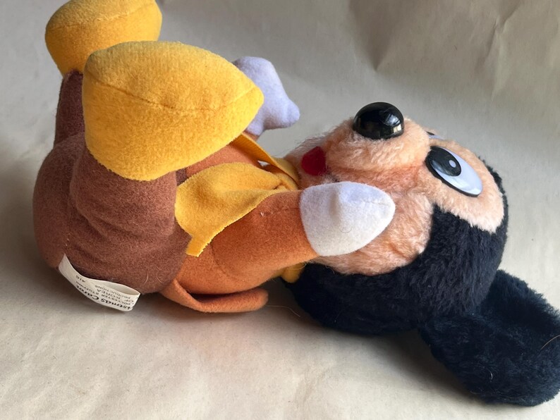 Op de afbeelding: Een pluche speelgoed met een stripfiguur met een zwarte kop, een oranje lichaam en gele accenten. Het speelgoed heeft een zwarte neus, grote ogen en een rode mond. Het speelgoed ligt op zijn zij.