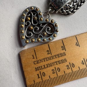 Pu&ograve; includere: Collana con pendente a forma di cuore color argento con dettagli dorati e intricate volute. Il cuore misura circa 2,5 cm di larghezza. La collana presenta una catena intrecciata e una chiusura decorativa.