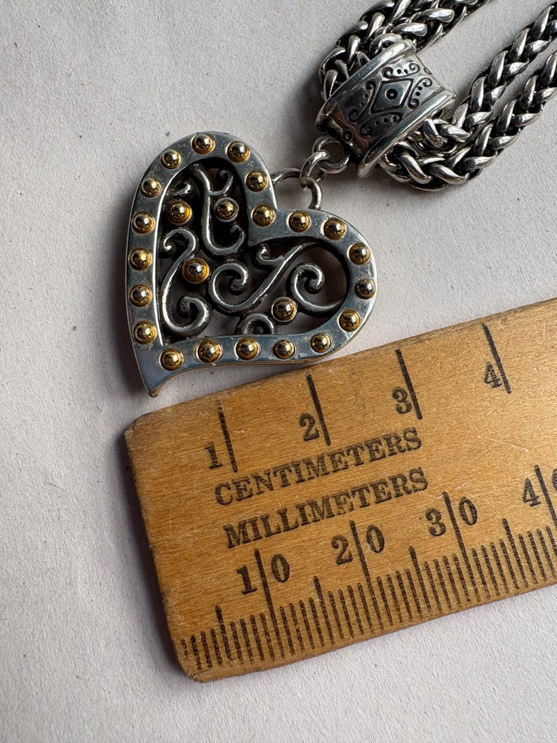 Pu&ograve; includere: Collana con pendente a forma di cuore color argento con dettagli dorati e intricate volute. Il cuore misura circa 2,5 cm di larghezza. La collana presenta una catena intrecciata e una chiusura decorativa.