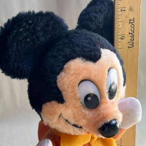 Op de afbeelding: Een vintage Mickey Mouse pluche speelgoed met zwarte oren, een perzikkleurig gezicht en een gele sjaal. Het pluche speelgoed houdt een houten liniaal vast die ongeveer 18 cm meet.
