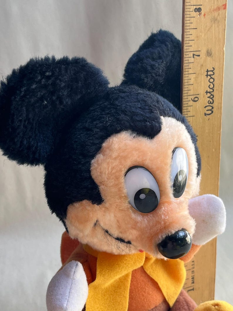 Op de afbeelding: Een vintage Mickey Mouse pluche speelgoed met zwarte oren, een perzikkleurig gezicht en een gele sjaal. Het pluche speelgoed houdt een houten liniaal vast die ongeveer 18 cm meet.