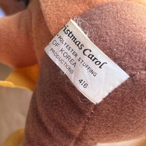 Op de afbeelding: Een close-up van een pluche speelgoed met een label met de tekst "Christmas Carol". Het speelgoed is voornamelijk bruin en bruin, met een gele accentkleur. Het label geeft ook "Polyester Stuffing of Korea Productions" en het nummer "416" aan.