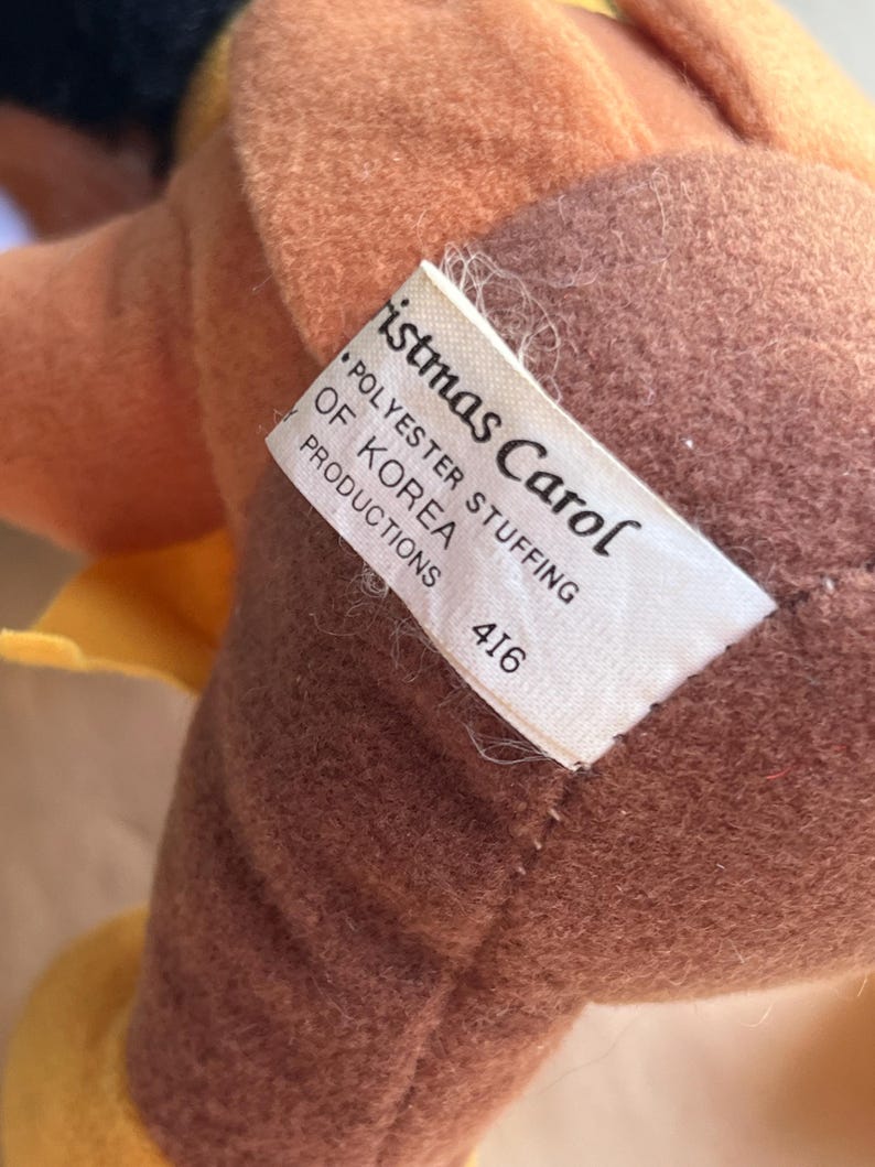 Op de afbeelding: Een close-up van een pluche speelgoed met een label met de tekst "Christmas Carol". Het speelgoed is voornamelijk bruin en bruin, met een gele accentkleur. Het label geeft ook "Polyester Stuffing of Korea Productions" en het nummer "416" aan.