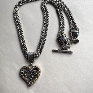 Pu&ograve; includere: Collana color argento con un ciondolo a forma di cuore. Il ciondolo presenta dettagli color oro e un design decorativo. La collana ha una catena intrecciata e una chiusura a toggle. Un gioiello elegante.