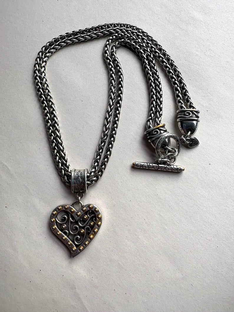 Pu&ograve; includere: Collana color argento con un ciondolo a forma di cuore. Il ciondolo presenta dettagli color oro e un design decorativo. La collana ha una catena intrecciata e una chiusura a toggle. Un gioiello elegante.