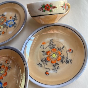 May include: A vintage teacup and saucers set. The cup and saucers are decorated with orange, blue, and white floral designs on a cream-colored background. The saucers have a light blue trim. The teacup has a yellow handle.