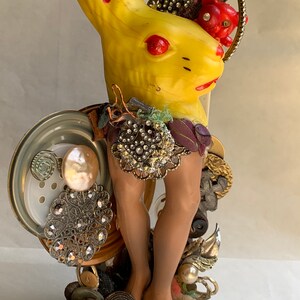 May include: A yellow plastic rabbit figurine with red accents, adorned with various vintage buttons, beads, and jewelry. The rabbit has brown legs and is standing on a base of assorted trinkets.