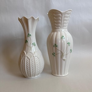 May include: Two white ceramic vases with scalloped rims and green shamrock motifs. The left vase features a textured diamond pattern on the lower body, while the right vase has vertical fluting and a leaf-like design on the neck.