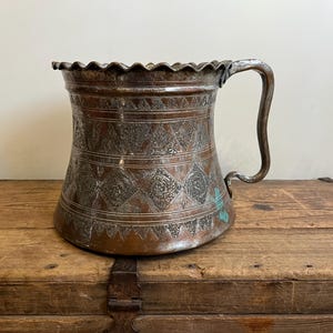 May include: An antique copper pitcher with an intricate embossed design. The pitcher has a wide mouth and a curved handle.