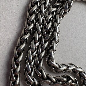 Pu&ograve; includere: Primo piano di una catena color argento con un motivo a spiga intrecciata. La catena &egrave; composta da maglie ad incastro, creando una superficie strutturata. Il metallo ha una qualit&agrave; lucida e riflettente, e la catena &egrave; esposta su uno sfondo grigio chiaro.