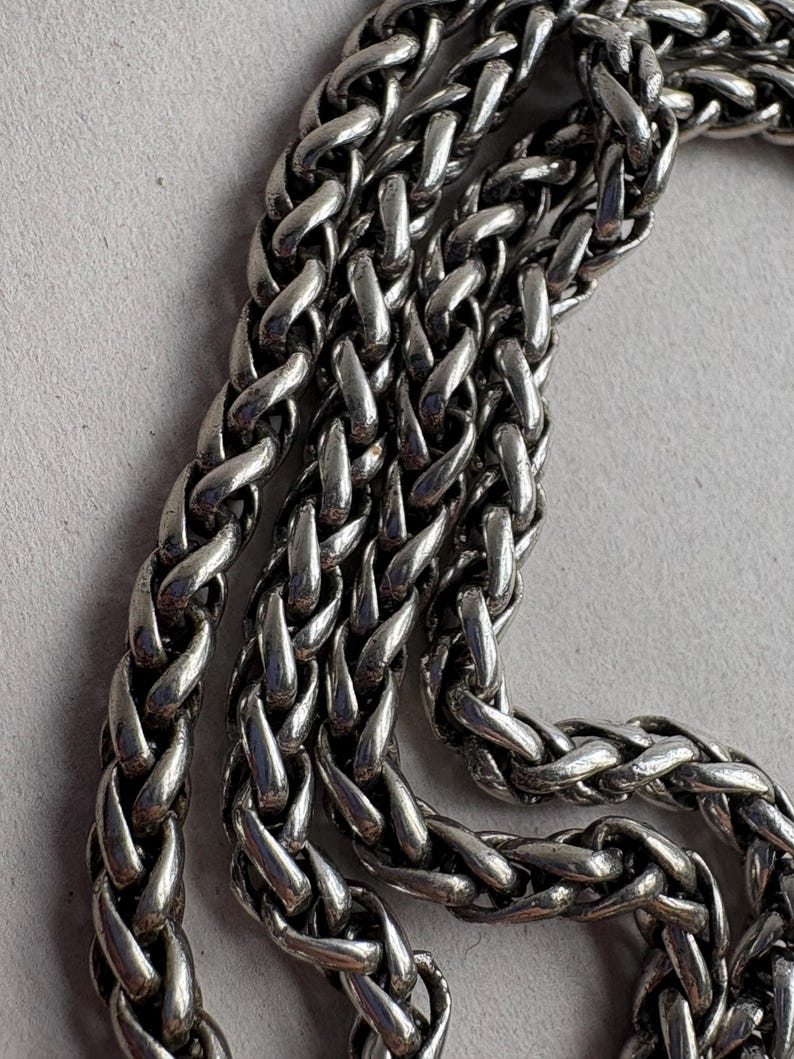 Pu&ograve; includere: Primo piano di una catena color argento con un motivo a spiga intrecciata. La catena &egrave; composta da maglie ad incastro, creando una superficie strutturata. Il metallo ha una qualit&agrave; lucida e riflettente, e la catena &egrave; esposta su uno sfondo grigio chiaro.