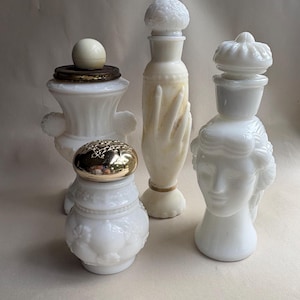 May include: A collection of four antique white glass perfume bottles. The bottles have unique shapes, including a hand, a woman's head, and a floral design. Some have gold or brass-colored lids. The bottles are displayed on a neutral background.