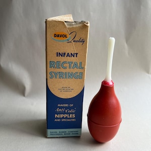 May include: Vintage infant rectal syringe with a red bulb and white tip, next to a cardboard box. The box reads "DAVOL Quality" and "INFANT RECTAL SYRINGE." The box also says "MADE IN UNITED STATES OF AMERICA."