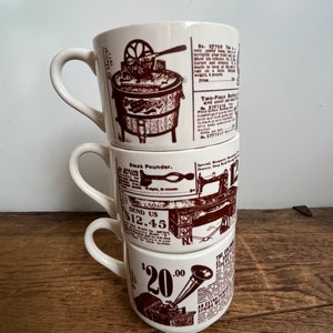 May include: A stack of three white ceramic mugs with brown illustrations of vintage advertisements. The top mug features a washing machine, the middle mug features a sewing machine, and the bottom mug features a gramophone. The text on the mugs reads "No. 27705", "Steak Pounder", "SEND US $12.45", and "$20.00".