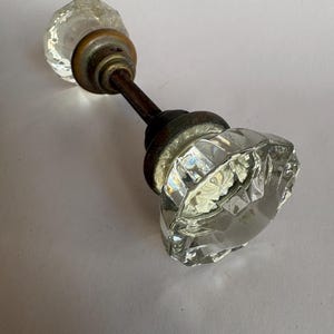 May include: A clear, faceted glass door knob set featuring two octagonal glass knobs connected by a central metal spindle. Each knob is attached to a circular, dark-toned metal base plate, showing signs of age and wear on the metallic components.