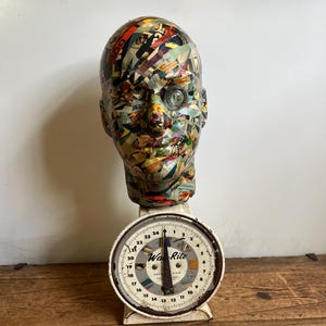 May include: A vintage scale with a head-shaped sculpture on top. The sculpture is covered in colorful paper fragments. The scale has a white face with black numbers and the text "Wa-Rite". The scale sits on a wooden surface.