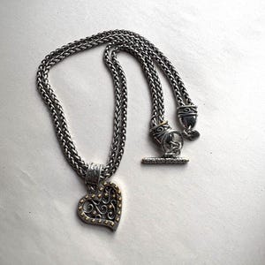 Pu&ograve; includere: Collana color argento con pendente a forma di cuore. Il pendente presenta un design a filigrana con dettagli dorati. La catena &egrave; a treccia con chiusura a toggle. Un gioiello elegante.