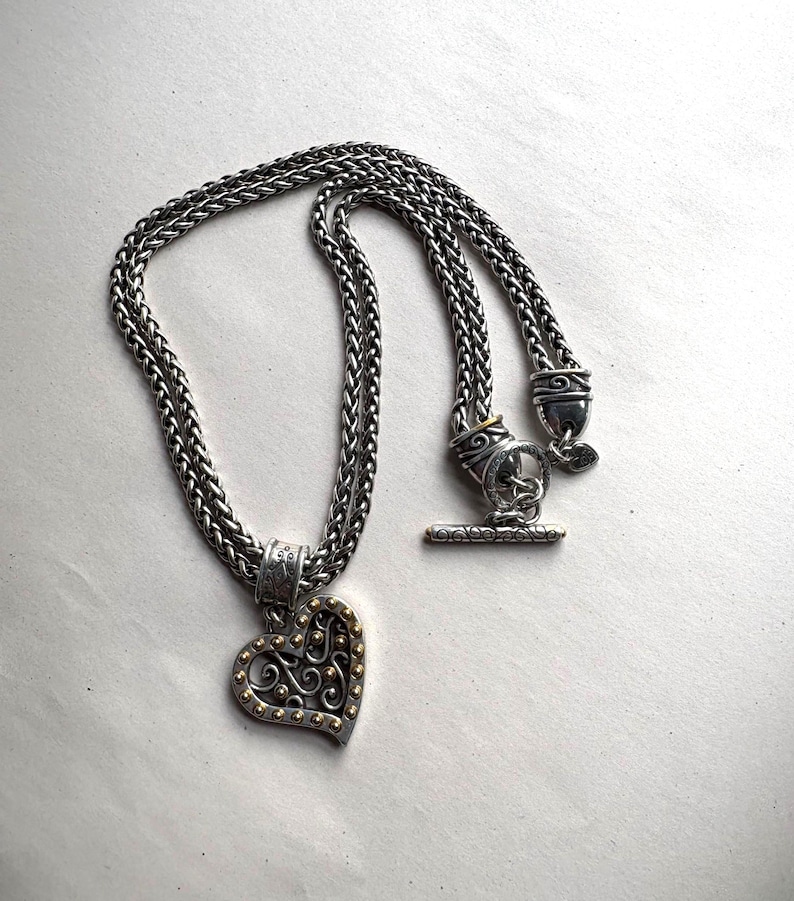 Pu&ograve; includere: Collana color argento con pendente a forma di cuore. Il pendente presenta un design a filigrana con dettagli dorati. La catena &egrave; a treccia con chiusura a toggle. Un gioiello elegante.