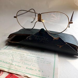 May include: Vintage gold-tone eyeglasses with a unique frame design, resting on a dark green leather case. Included are several vintage documents, including a certificate guaranteeing the lenses for life from Raynard Optical Co.