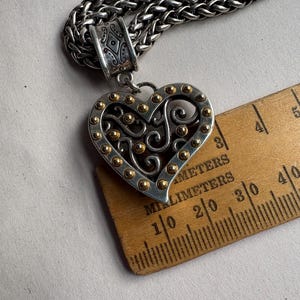 Pu&ograve; includere: Collana con pendente a forma di cuore color argento con dettagli dorati. Il pendente a forma di cuore presenta un design traforato ed &egrave; attaccato a una catena spessa e strutturata. Il pendente &egrave; largo circa 4 cm.