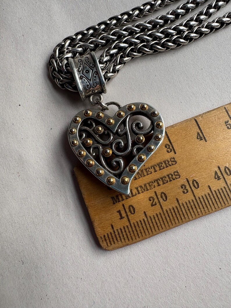 Pu&ograve; includere: Collana con pendente a forma di cuore color argento con dettagli dorati. Il pendente a forma di cuore presenta un design traforato ed &egrave; attaccato a una catena spessa e strutturata. Il pendente &egrave; largo circa 4 cm.