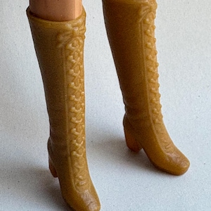 May include: A pair of vintage, knee-high, tan-colored boots. The boots have a textured, decorative pattern along the sides and a small heel. The boots are made of a hard plastic material.