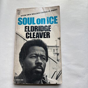 May include: A paperback book cover with a black and white photo of Eldridge Cleaver. The title of the book is "Soul on Ice" and the author is Eldridge Cleaver. The book cover is white with blue text and a black and white photo.