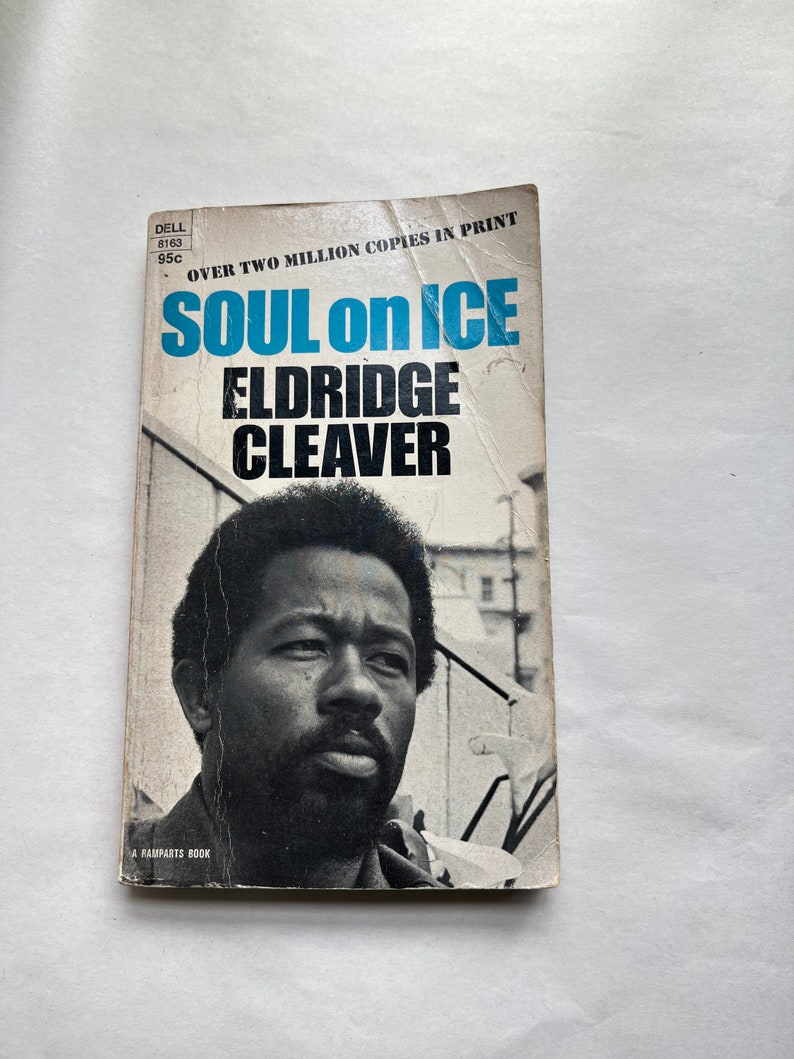 May include: A paperback book cover with a black and white photo of Eldridge Cleaver. The title of the book is "Soul on Ice" and the author is Eldridge Cleaver. The book cover is white with blue text and a black and white photo.