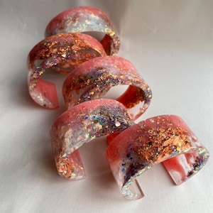May include: Five open-ended resin bracelets. Each bracelet is a translucent pink with embedded gold leaf and iridescent glitter. The bracelets are stacked, showing the open cuff design.