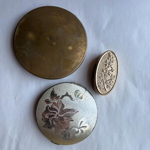 May include: An assortment of vintage compacts and a small pillbox. The compacts are round, one is gold-toned, and the other is silver with a floral design. The pillbox is oval and gold-toned, featuring an embossed floral pattern. These items are likely from the mid-20th century.