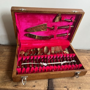 May include: A wooden box with a red velvet lining contains a set of gold-toned cutlery. The cutlery includes forks, spoons, and knives, all with wooden handles. The box has a brass latch and hinges.