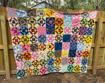 May include: A colorful patchwork quilt with a variety of patterned squares in shades of pink, blue, yellow, and brown. The quilt is displayed outdoors, draped over a wooden fence, showcasing its handmade quality and vibrant design.
