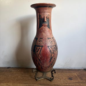 May include: A tall, terracotta-colored ceramic vase featuring a sculpted human face on the neck and intricate black geometric patterns on the body. The vase is supported by a decorative, three-legged brass-toned metal stand with ornate scrollwork.