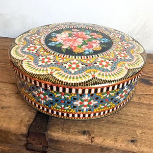 May include: Round, vintage tin with a floral design on the lid. The lid features a black center with pink and yellow flowers. The sides and lid are decorated with colorful geometric patterns in red, blue, yellow, and white. The tin sits on a wooden surface.