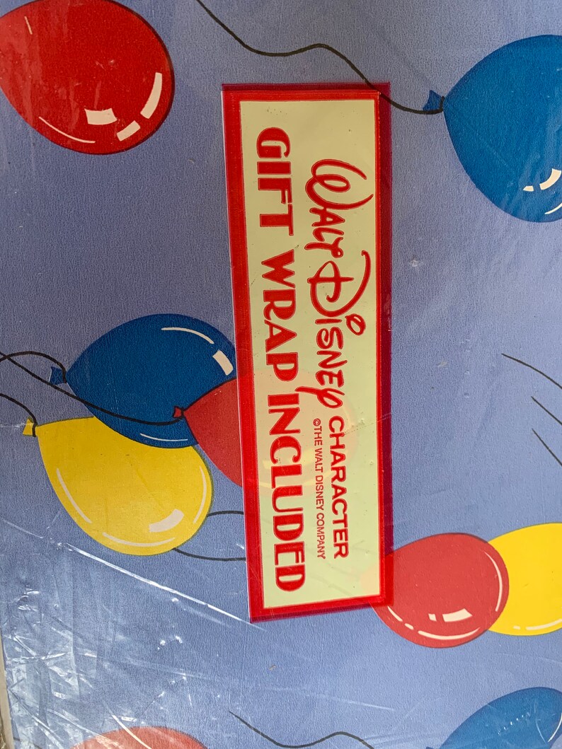 May include: Blue wrapping paper with a red border and a red and white label that says "Walt Disney Character Gift Wrap Included" with a copyright symbol and "The Walt Disney Company". The paper is decorated with colourful balloons.