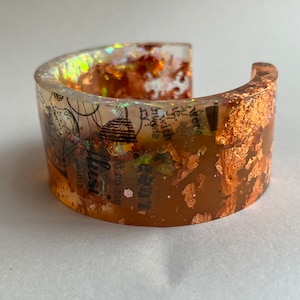 May include: A clear resin cuff bracelet with copper leaf accents and a vintage map design. The map includes the text "2023" and "illus."