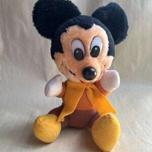 Op de afbeelding: Een pluche Mickey Mouse speelgoed met zwarte oren, een bruin gezicht en een zwarte neus. Het speelgoed draagt een oranje shirt, gele schoenen en een gele sjaal. Het speelgoed zit met uitgestrekte armen.