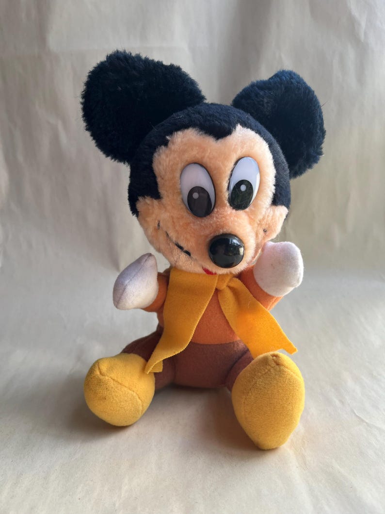 Op de afbeelding: Een pluche Mickey Mouse speelgoed met zwarte oren, een bruin gezicht en een zwarte neus. Het speelgoed draagt een oranje shirt, gele schoenen en een gele sjaal. Het speelgoed zit met uitgestrekte armen.