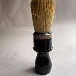 May include: A black shaving brush with a silver band and a handle that reads "OMEGA ITALY". The brush has a dense, light brown bristle head. The handle is glossy and has a rounded shape.
