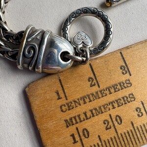 Pu&ograve; includere: Bracciale argentato con chiusura decorativa. Il bracciale presenta un design a catena dettagliato e una chiusura a levetta. La chiusura include un anello decorativo con un ciondolo a forma di cuore. Un righello di legno con segni in centimetri e millimetri &egrave; nell'immagine.