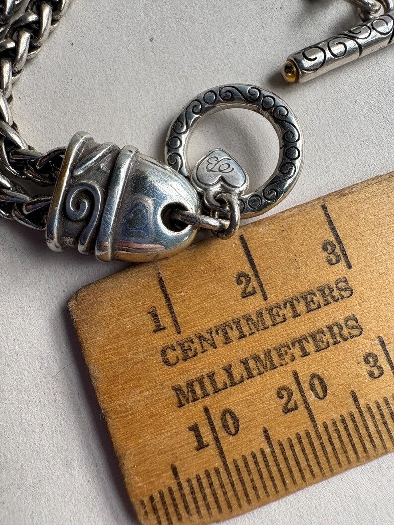 Pu&ograve; includere: Bracciale argentato con chiusura decorativa. Il bracciale presenta un design a catena dettagliato e una chiusura a levetta. La chiusura include un anello decorativo con un ciondolo a forma di cuore. Un righello di legno con segni in centimetri e millimetri &egrave; nell'immagine.