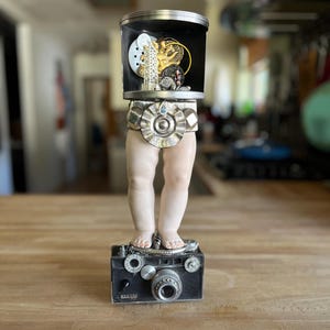 May include: A unique art piece featuring a vintage camera base supporting a pair of porcelain legs. Above, a cylindrical container holds miniature sculptures. The piece is set on a wooden surface.