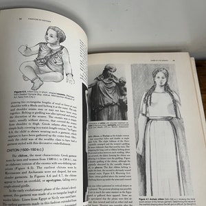 May include: A black and white illustration of a woman wearing a long, flowing garment called a chiton. The chiton is belted at the waist and has a draped overfold. The illustration is from a book about fashion history.